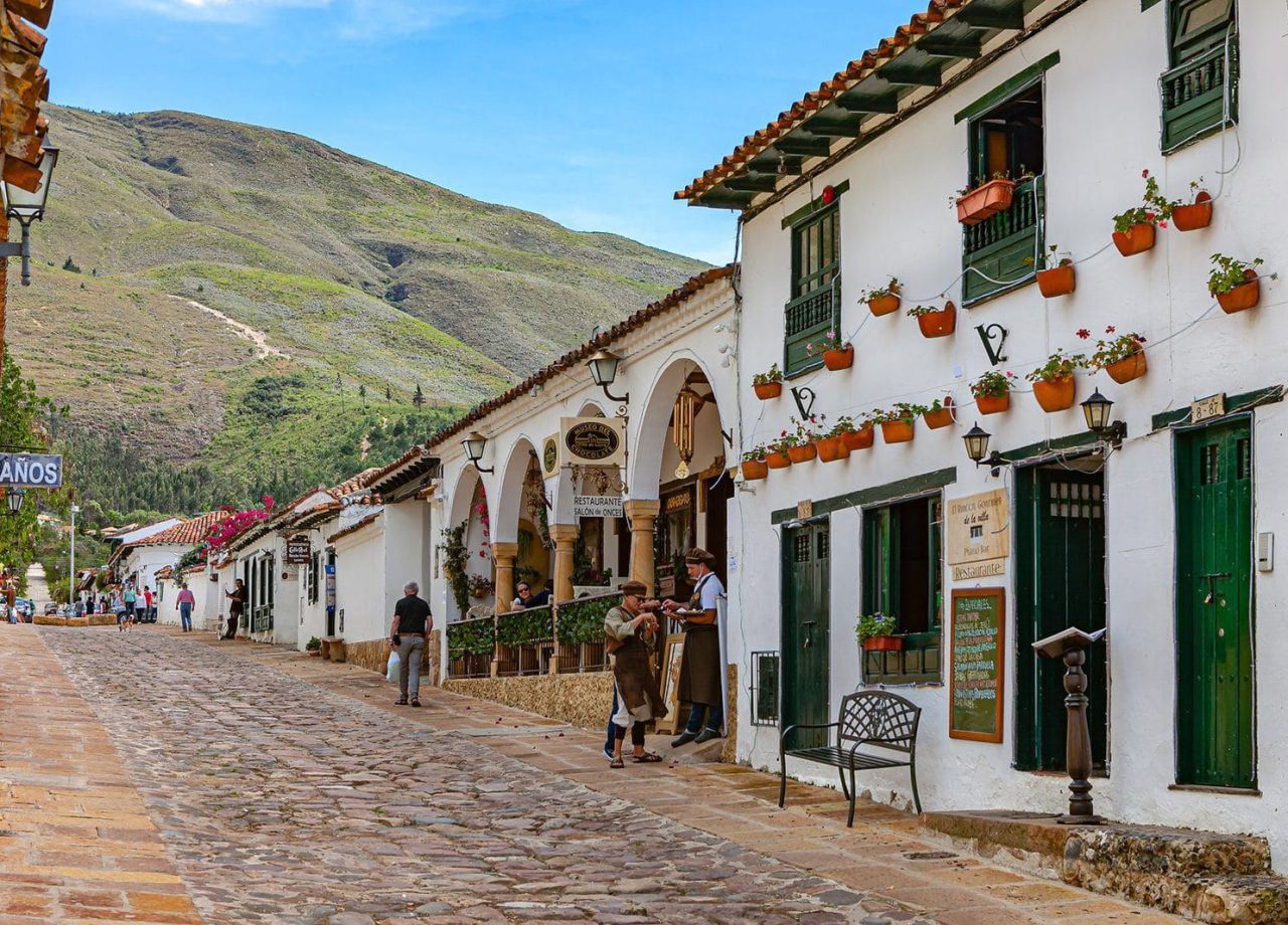 Villa de Leyva, Colombia - 26 de noviembre de 2017: buscando Calle 13 en la histórica ciudad colonial del siglo XVI de Villa de Leyva en el departamento de Boyacá. A la derecha hay una fila de restaurantes uno tras otro, con un miembro del personal parado en la calle tratando de atraer a los clientes a sus restaurantes. En el fondo lejano hay una sección de la Cordillera de los Andes. Fundada en 1572 y ubicada a poco más de 7000 pies sobre el nivel medio del mar en la Cordillera de los Andes, Villa de Leyva fue declarada Monumento Nacional por el gobierno colombiano en 1954 para proteger su arquitectura y patrimonio colonial. The Town también fue uno de los lugares para la película Cobra Verde de Werner Herzog y la telenovela Zorro en español. Foto tomada a la luz del sol del mediodía; formato horizontal. Copie el espacio.